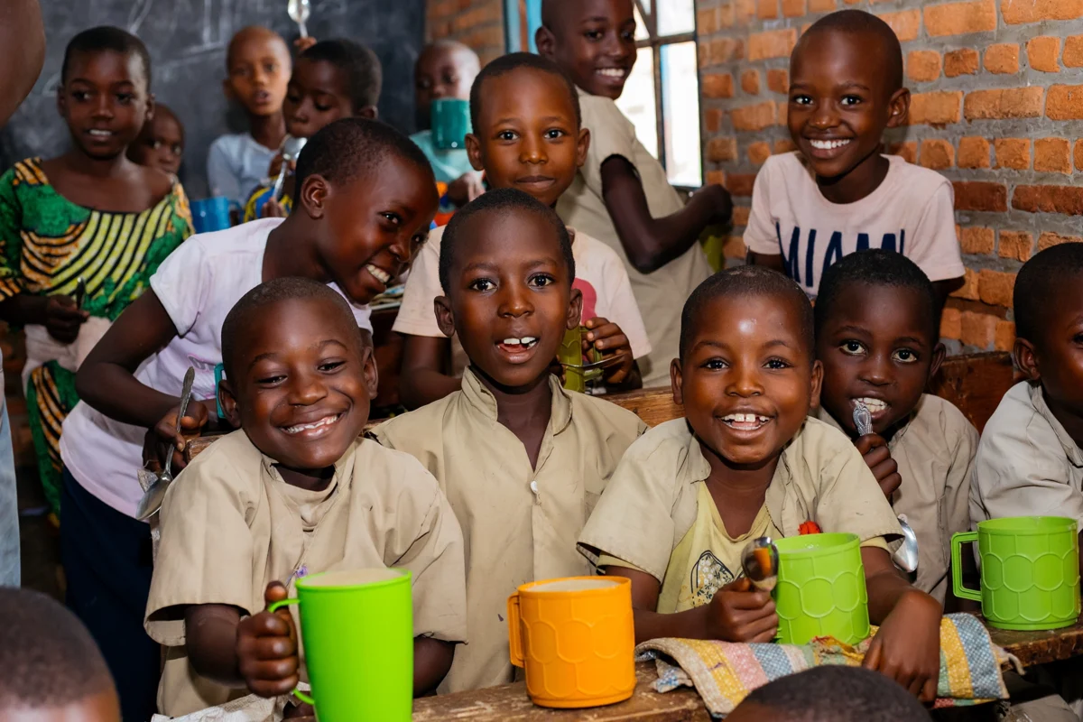 Lachende Kinder in Burundi essen gemeinsam Porridge in der Schule - das Projekt „Ein Becher am Tag“ von Helfende Hände verbessert Gesundheit und Konzentration durch tägliche Schulmahlzeiten.