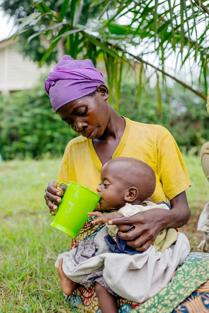 Mutter in Burundi füttert ihr geschwächtes Kind mit Porridge aus einem Becher - das Ernährungsprogramm von Helfende Hände schützt vor Mangelernährung und gibt Familien Hoffnung.