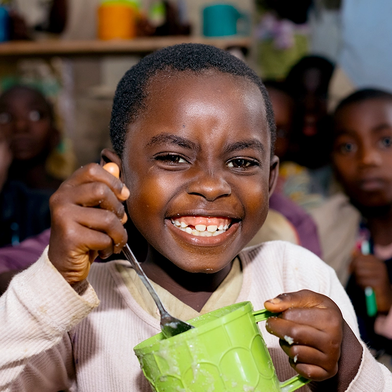 Glückliches Kind in Burundi isst Porridge aus einem Becher - das Hilfsprojekt „Ein Becher am Tag“ von Helfende Hände schenkt Kindern neue Kraft, Gesundheit und Schulchancen.