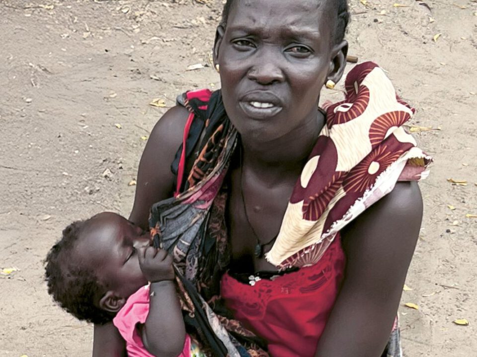 Mutter im Südsudan sitzt erschöpft auf dem Boden und wiegt ihr unterernährtes Kind im Arm. In der staubigen, kargen Umgebung wird die humanitäre Notlage deutlich.