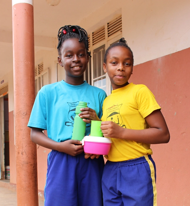 Zwei Schülerinnen der Zana Community Christian School in Uganda mit Trinkflaschen und Essensbox – Schulprojekt für Bildung und gesunde Ernährung.