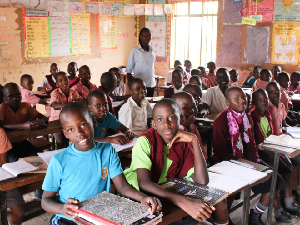 helfende hände - Ein Klassenzimmer voller Schüler, die an ihren Schreibtischen sitzen, nach vorne blicken und ihre Hefte aufgeschlagen haben. Der Lehrer steht hinten im Raum. An den Wänden hängen bunte Lehrplakate. helfende hände ggmbh hamburg