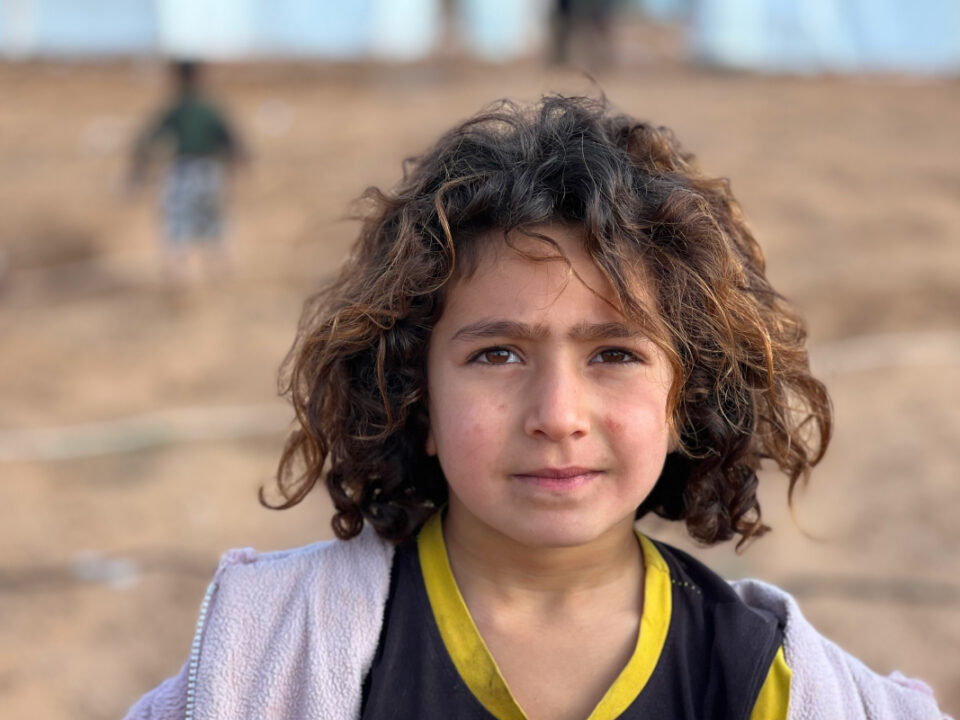 helfende hände - Ein Kind mit lockigem braunem Haar und ernstem Gesichtsausdruck steht draußen auf sandigem Boden; es trägt ein schwarz-gelbes Hemd und eine helle Jacke. Der Hintergrund ist unscharf, im Hintergrund ist eine weitere Person zu erkennen. helfende hände ggmbh hamburg