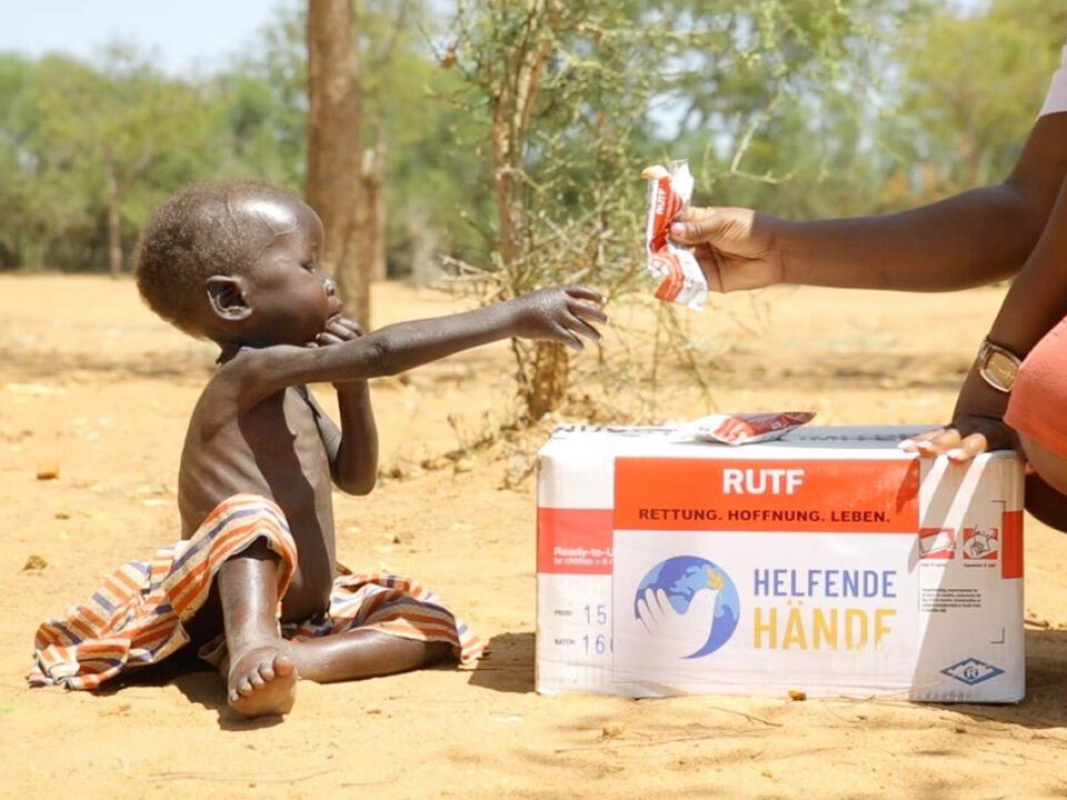 helfende hände - Ein unterernährtes Kind sitzt auf trockenem Boden und streckt die Hand nach einem Energieriegel aus, den ihm ein Erwachsener reicht. Neben dem Kind steht eine weiße Schachtel mit der Aufschrift „RUTF“ und „Helfende Hände“. Im Hintergrund sind vereinzelte Bäume und trockene Erde zu sehen. helfende hände ggmbh hamburg