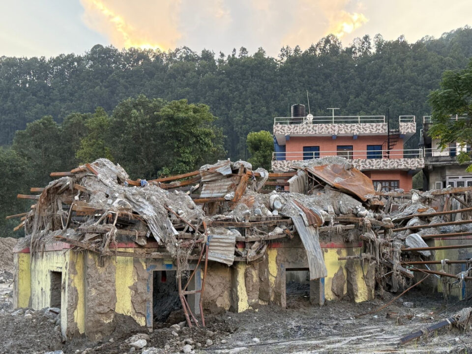 helfende hände - Ein gelbes Gebäude, das durch Trümmer schwer beschädigt wurde, im Hintergrund ein noch stehendes zweistöckiges Backsteinhaus, dokumentiert von Nothilfe Nepal. helfende hände ggmbh hamburg